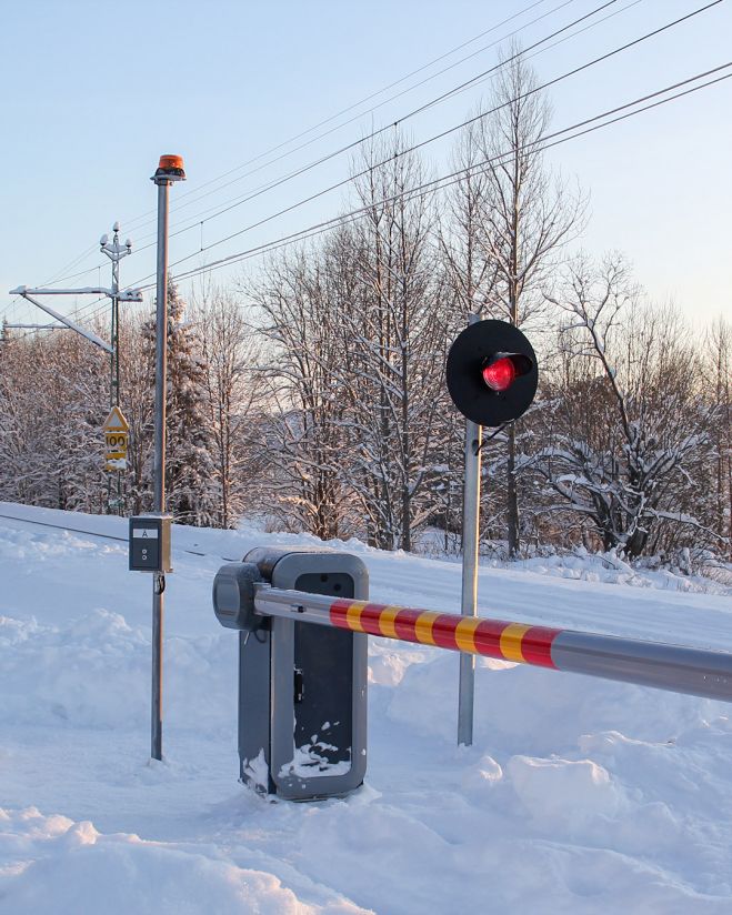 Snowy railroad crossing with a lowered barrier and red-light signal on the side. Snow-covered trees and tracks are visible.
